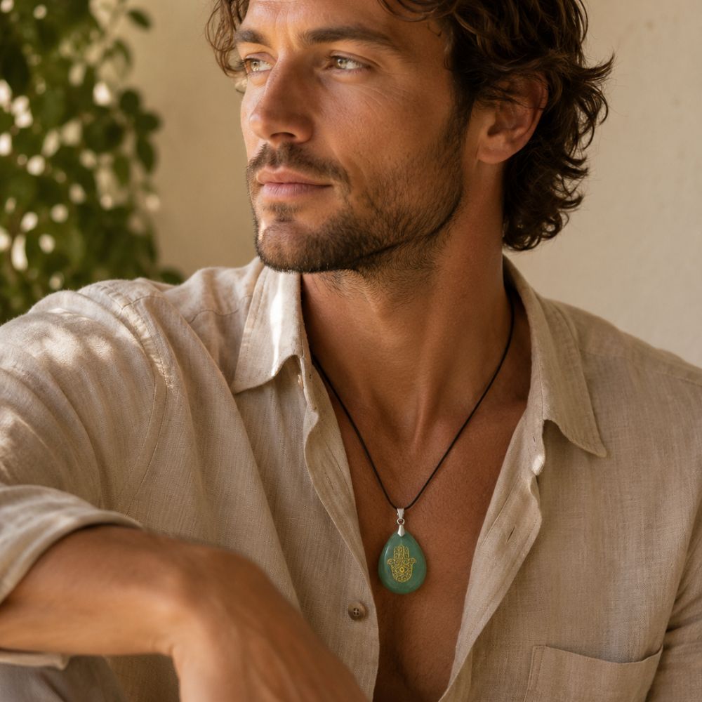 A man wearing a light beige linen shirt and a black cord necklace with a round turquoise pendant featuring a gold hamsa hand design, set against a warm, softly lit background.