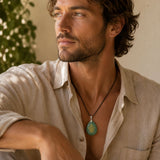 A man wearing a light beige linen shirt and a black cord necklace with a round turquoise pendant featuring a gold hamsa hand design, set against a warm, softly lit background.