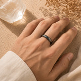 A close-up of a person's hand wearing a Hematite ring, resting on a textured beige surface with dried flowers and a clear crystal nearby.