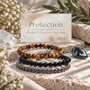 Three beaded protection bracelets displayed on a stone surface with a card reading 'Protection' and 'Earth Elements' branding, surrounded by dried flowers, greenery, and crystals. The bracelets are Tiger's Eye, Black Obsidian, and Hematite.