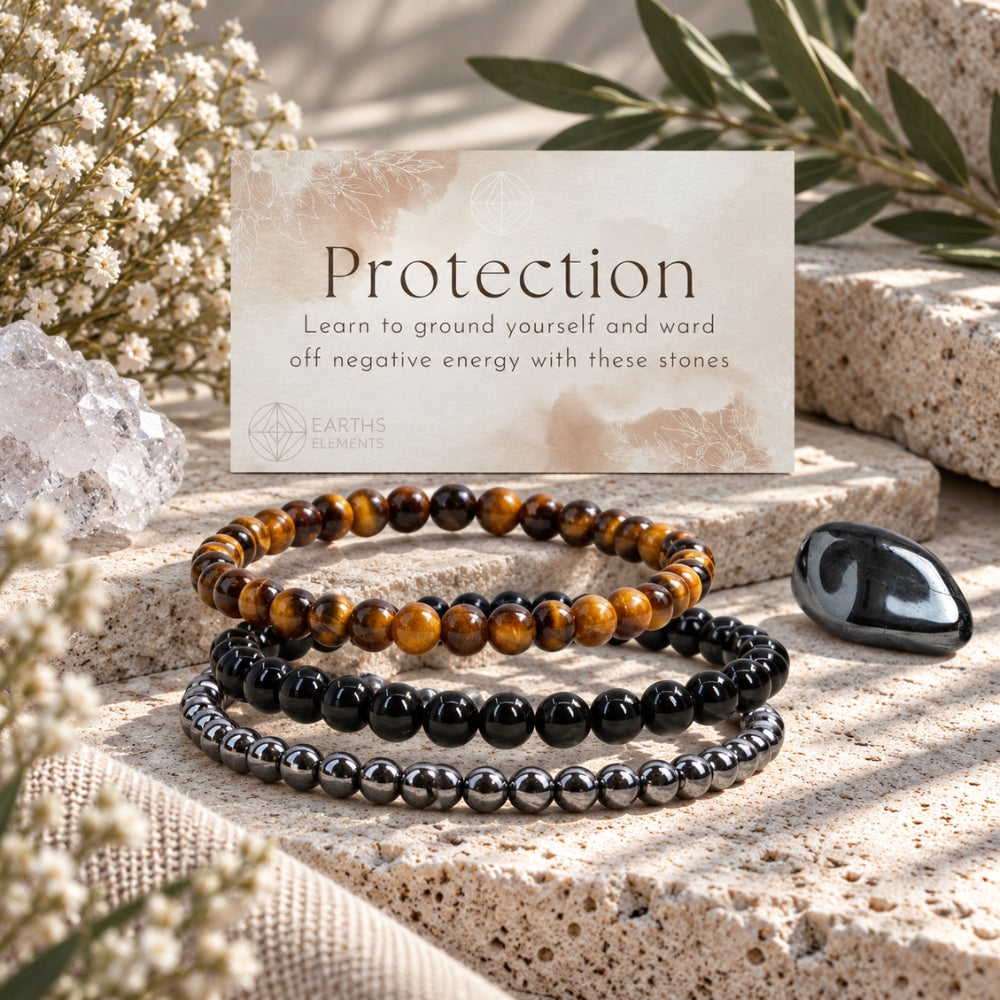 Three beaded protection bracelets displayed on a stone surface with a card reading 'Protection' and 'Earth Elements' branding, surrounded by dried flowers, greenery, and crystals. The bracelets are Tiger's Eye, Black Obsidian, and Hematite.