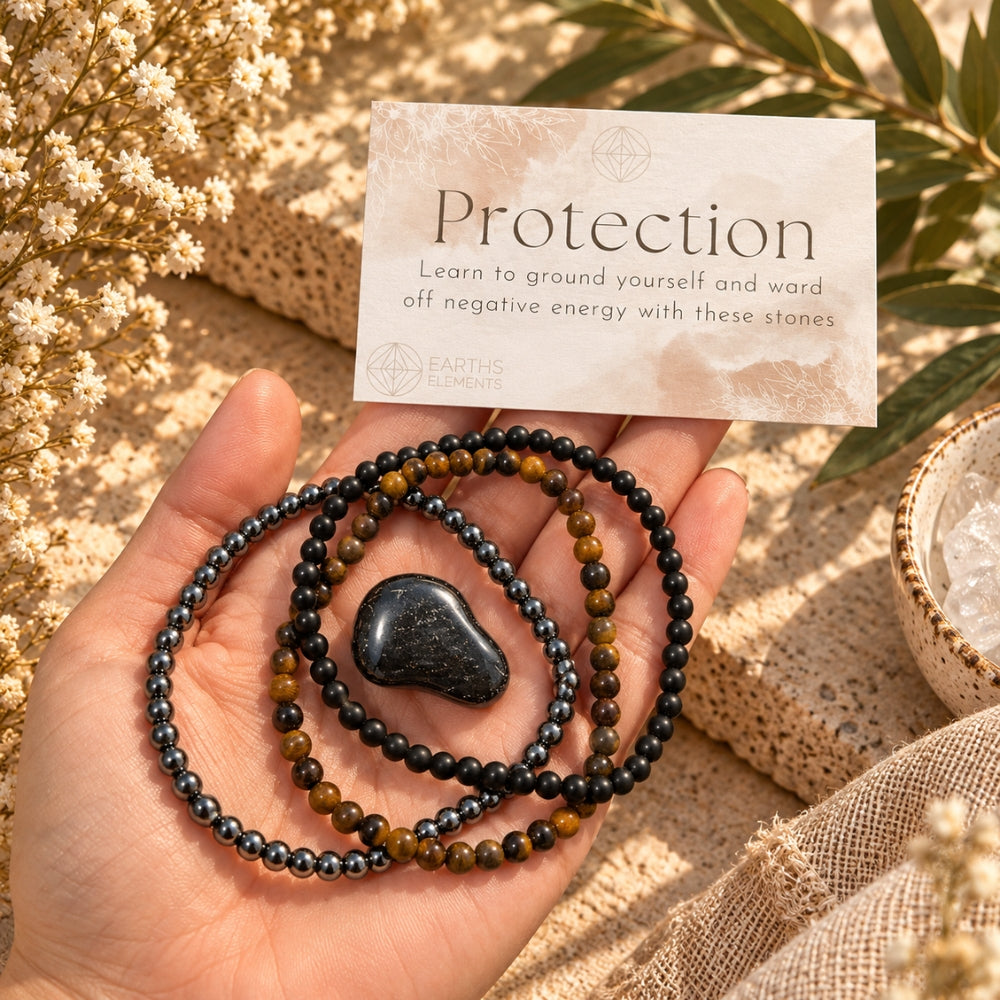 A hand holds a set of protective crystal bead bracelets and a tumbled black stone, with an 'Earth Energy' card labeled 'Protection' in the background. The bracelets are Tiger's Eye, Black Obsidian, and Hematite.