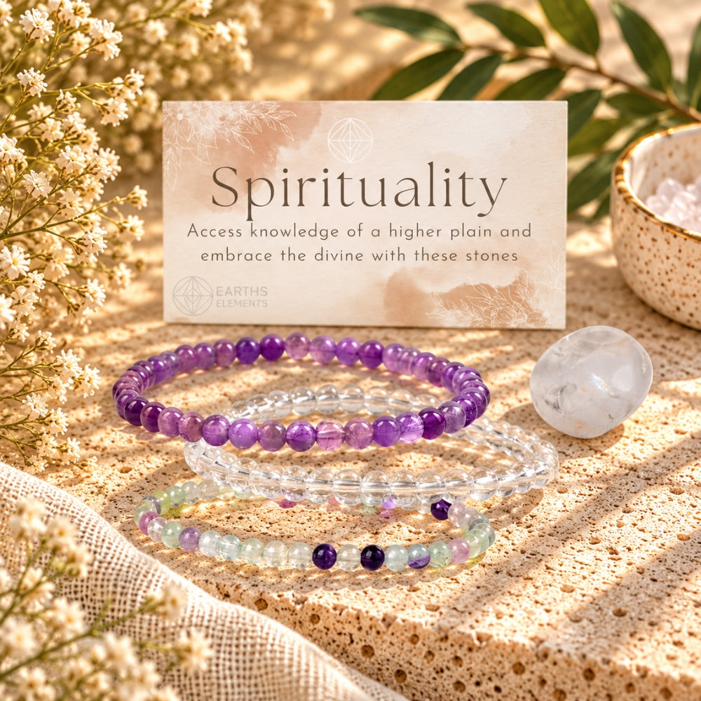 Three beaded stretch bracelets displayed on a textured beige surface with dried flowers, green leaves, and a card reading 'Spirituality' by Earths Elements. The bracelets include Amethyst, clear quartz, and fluorite.