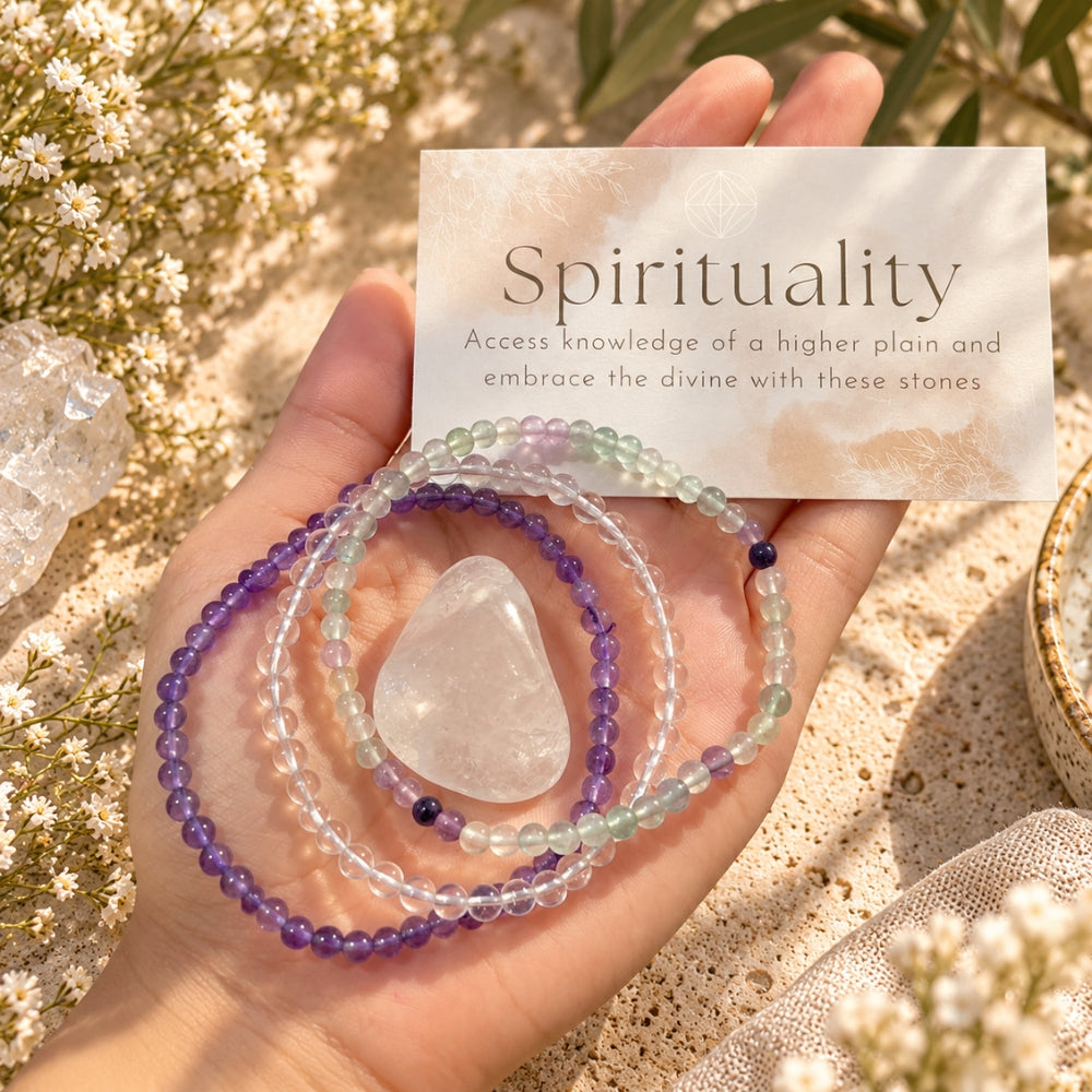 A hand holds a set of three beaded spiritual bracelets that are Amethyst, clear quartz, and fluorite with a clear tumbled stone, surrounded by small white flowers and a card that reads 'Spirituality'.