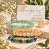 Three beaded gemstone bracelets arranged on a stone slab with a card reading 'Abundance' behind them. The bracelets are green aventurine, citrine, and moss agate.