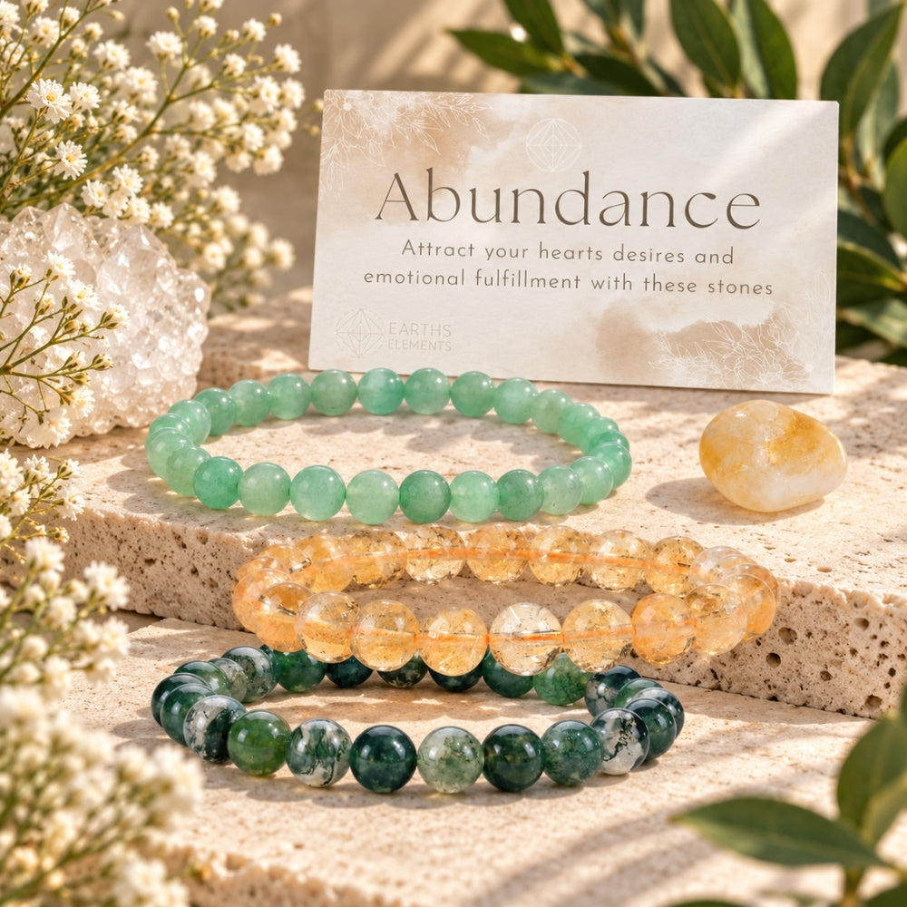 Three beaded gemstone bracelets arranged on a stone slab with a card reading 'Abundance' behind them. The bracelets are green aventurine, citrine, and moss agate.