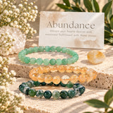 Three beaded gemstone bracelets arranged on a stone slab with a card reading 'Abundance' behind them. The bracelets are green aventurine, citrine, and moss agate.