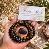 A hand holds a set of protective crystal bead bracelets and a tumbled black stone, with an 'Earth Energy' card labeled 'Protection' in the background. The bracelets are Tiger's Eye, Black Obsidian, and Hematite.