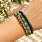Three beaded stretch bracelets worn on a wrist: moss agate, black onyx, and tiger's eye, shown against a softly blurred outdoor background with small white flowers.