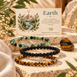 Three beaded stretch bracelets arranged on a stone surface in front of a printed card that reads "Earth." The bracelets are made of moss agate, black onyx, and tiger's eye.