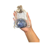 A hand holding a clear drawstring pouch with a beige burlap-style top, containing a blue beaded bracelet and a small card labeled Moon.