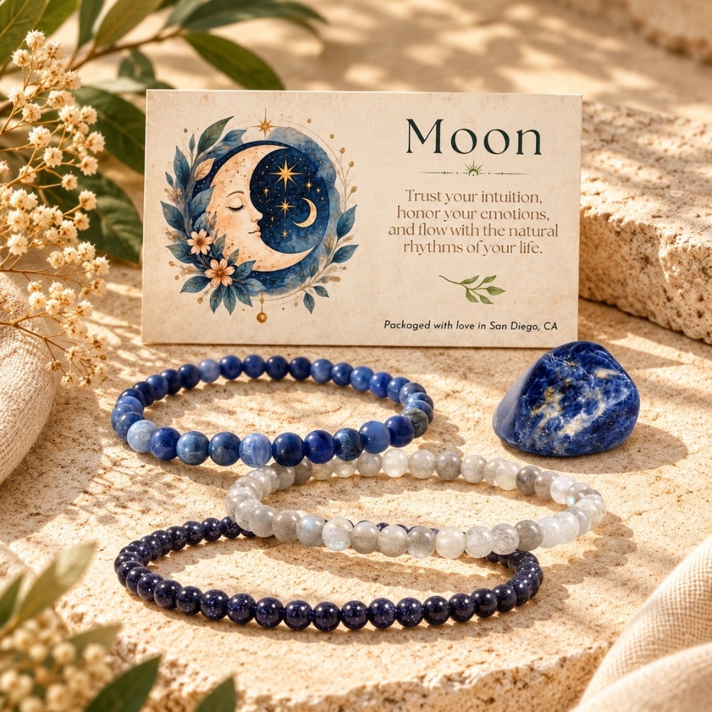 Three beaded stretch bracelets in shades of navy, light blue-gray, and deep navy are arranged on a sandy surface beside a blue stone and a printed card featuring a moon illustration and the word 'Moon'.