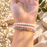 Three beaded stretch bracelets in howlite, labradorite, and rose quartz, shown stacked on a person's wrist with a warm, sunlit background of dried flowers and textured stones.