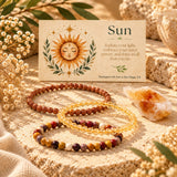 Three beaded stretch bracelets arranged on a textured stone surface beside a printed card labeled "Sun" with a sun illustration. The bracelets includeCitrine, Goldstone, Mookaite and a crystal citrine cluster around them.