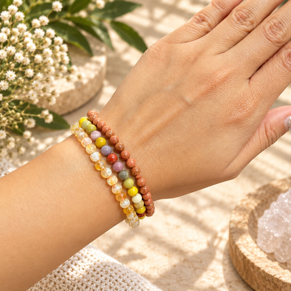A close-up of a person's wrist wearing three beaded stretch bracelets in warm earthy and pastel tones, with a hand resting above it against a softly styled background with flowers and crystals.