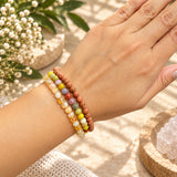 A close-up of a person's wrist wearing three beaded stretch bracelets in warm earthy and pastel tones, with a hand resting above it against a softly styled background with flowers and crystals.