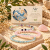 Three beaded stretch bracelets in pastel pink, light blue, and lavender purple are arranged on a textured stone surface in front of a cream card labeled 'Air' with a feather illustration. The scene includes dried flowers, green leaves, and a rose quartz crystal against a warm natural background.