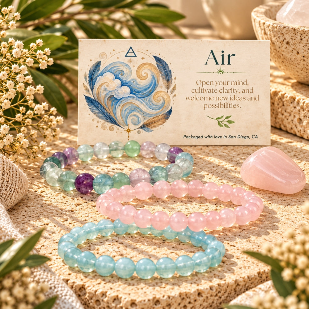 Three beaded stretch bracelets in fluorite, rose quartz, and opalite are arranged on a textured beige surface beside a printed card labeled 'Air' with a feather illustration. The scene includes decorative flowers, burlap, and a rose quartz crystal in a warm styled setting.