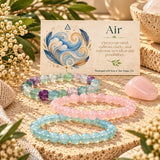 Three beaded stretch bracelets in fluorite, rose quartz, and opalite are arranged on a textured beige surface beside a printed card labeled 'Air' with a feather illustration. The scene includes decorative flowers, burlap, and a rose quartz crystal in a warm styled setting.