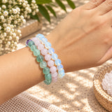 Three beaded stretch bracelets worn on a person's wrist, featuring fluorite, rose quartz, and opalite bracelets. The background is a warm, sunlit setting with dried flowers and a woven mat.