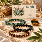 Three beaded stretch bracelets arranged on a stone surface with a small printed card behind them and a tiger's eye stone nearby. The bracelets are made of round beads they are moss agate, black obsidian, and tiger's eye.