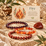 Three beaded stretch bracelets in deep red, orange, and translucent amber tones are arranged on a textured stone surface beside a printed card labeled 'Fire' with a flame illustration. The scene includes decorative dried flowers, leaves, and a polished red stone in a warm styled setting.