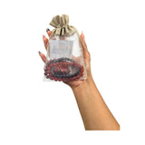 A hand holding a clear drawstring pouch with a burlap-style top, containing a red and dark gray beaded bracelet set with a central carved stone, against a plain white background.