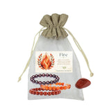 A fire-themed gemstone bracelet set in a sheer drawstring pouch with a flame illustration label. The set includes three beaded braceletsCarnelian, Garnet, and Red Jasper, along with a red jasper tumbled stone, all arranged in front of a light gray fabric pouch on a white background.