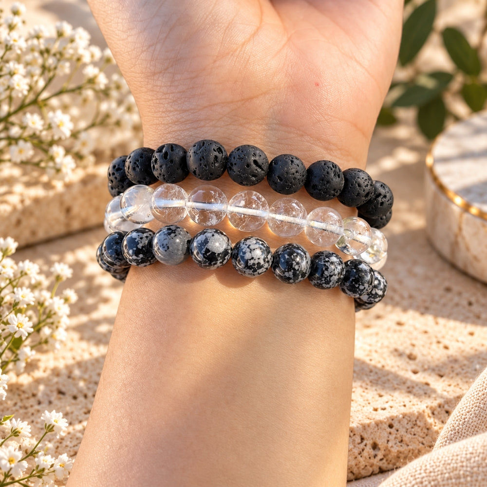 Three beaded stretch bracelets worn on a person's wrist. The top bracelet is made of dark black lava stones, the middle bracelet is made of clear quartz beads, and the bottom bracelet is made of snowflake obsidian.