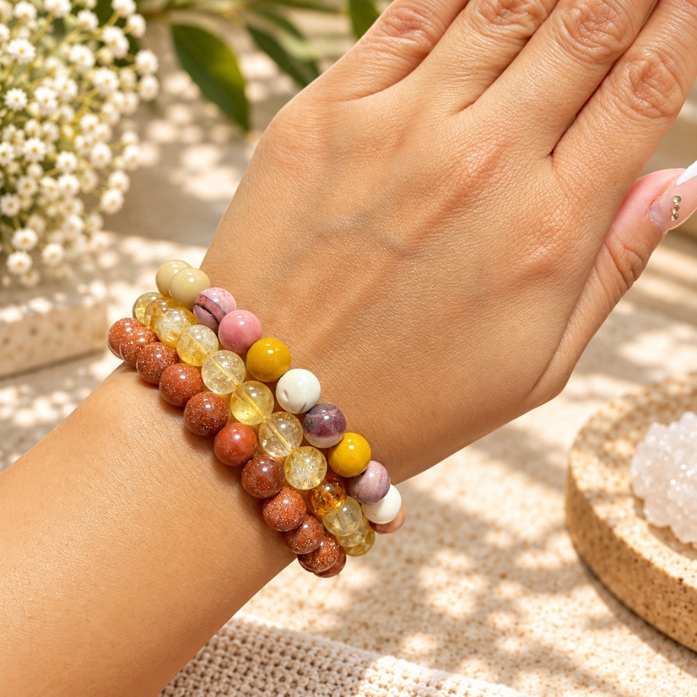 A close-up of a person's wrist wearing goldstone, citrine, and mookaite bracelets made of multicolored gemstone-like beads, photographed outdoors with soft sunlight and blurred flowers in the background.