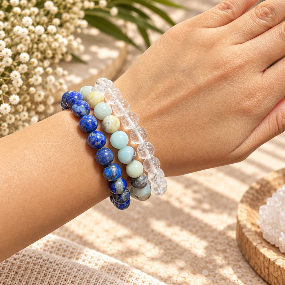 A close-up of a person's wrist wearing three beaded stretch bracelets: a blue lapis lazuli bracelet, an Amazonite and clear quartz bracelet. The background features a neutral textured surface with soft-focus white flowers.