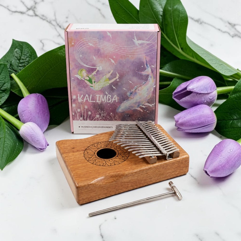 A kalimba thumb piano with a wooden body, metal tines, and a metal wrench, styled on a white marble surface with purple tulips and green leaves, with its illustrated pink and purple box behind it.