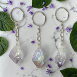 Three Aura Amethyst keychains on a marble surface with purple flowers and green leaves.