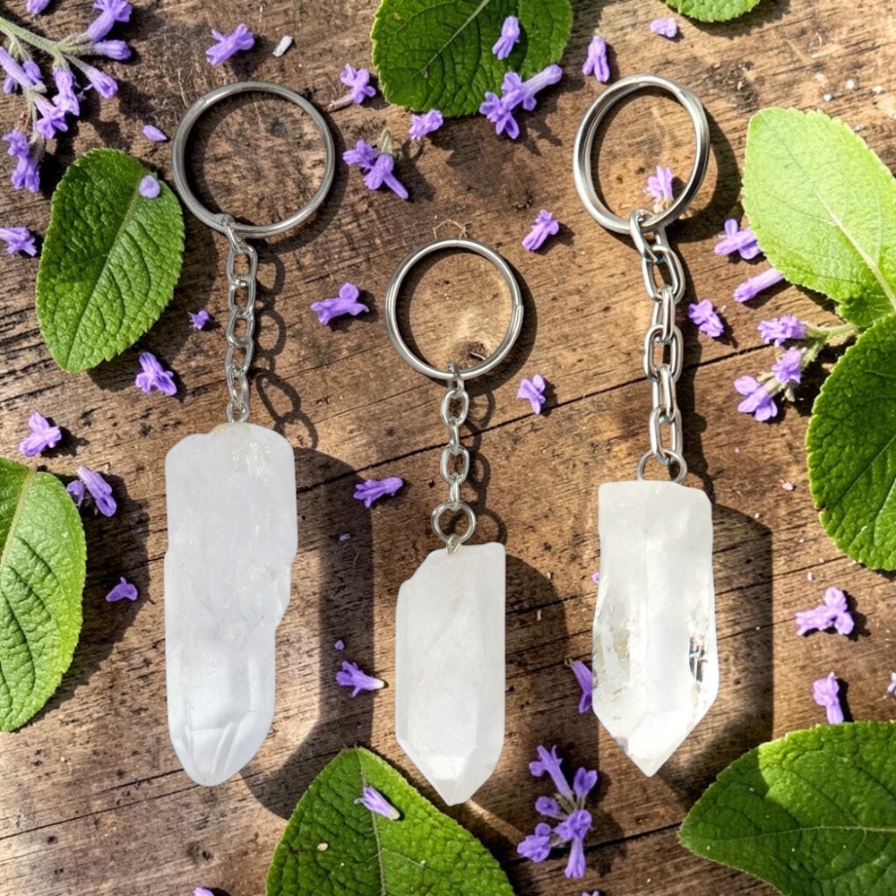 Three Clear Quartz keychains on a wooden surface with purple flowers and green leaves.