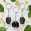 Three black Obsidian keychains on a marble surface with green leaves and yellow flowers.