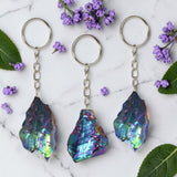 Three aura keychains on a marble surface with purple flowers and green leaves.