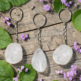 Three Clear Quartz keychains on a wooden surface with green leaves and purple flowers.