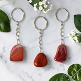 Three Red Jasper keychains on a marble surface with green leaves and white flowers.