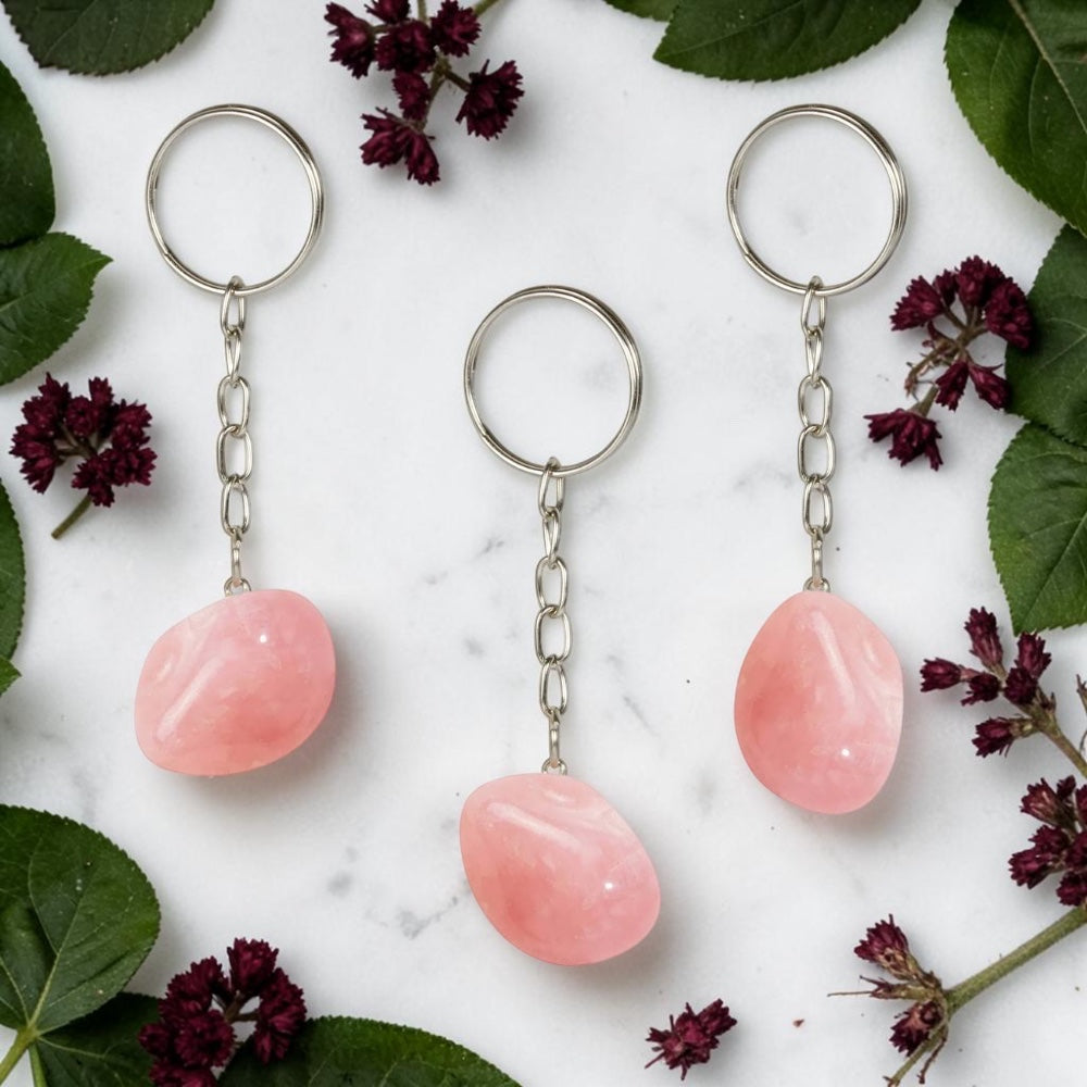 Three Rose Quartz keychains on a marble surface with green leaves and purple flowers.