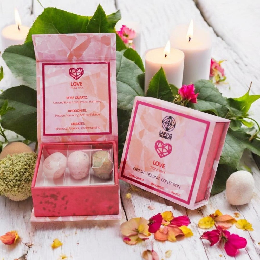 Pink packaging with tumbled crystals and candles on a wooden surface with flowers and leaves.