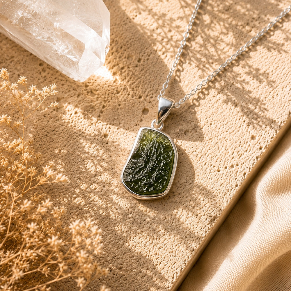 Moldavite Crystal Necklace with Chain - 925 displayed on a textured beige surface with warm sunlight, a clear crystal, and dried flowers.