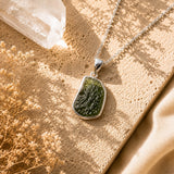 Moldavite Crystal Necklace with Chain - 925 displayed on a textured beige surface with warm sunlight, a clear crystal, and dried flowers.