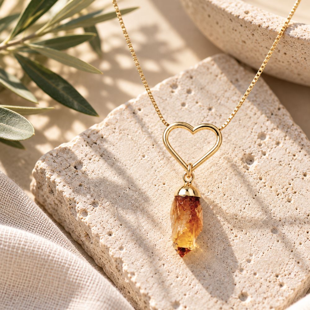 Gold heart necklace with a dangling raw citrine crystal pendant displayed on natural stone in soft sunlight, Earths Elements jewelry.