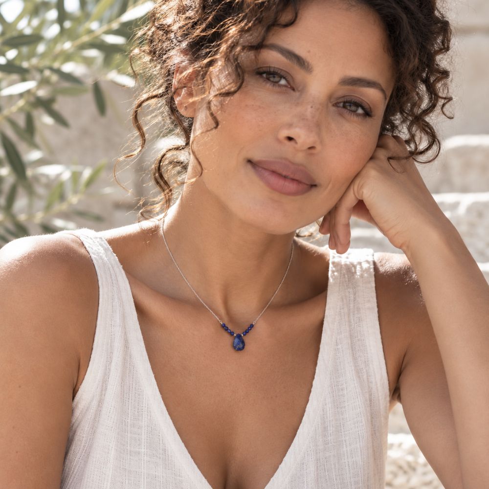 Woman with naturally curly dark hair wearing a delicate Lapis Lazuli Beaded Necklace with deep blue gemstone beads, a faceted teardrop pendant, and a fine silver chain, styled with a sleeveless ivory linen top.