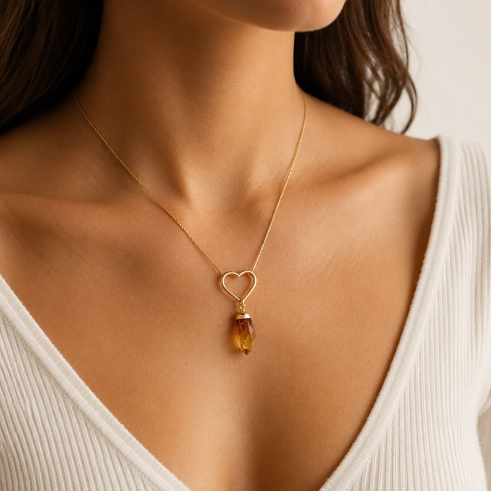 A close-up of a woman wearing a delicate gold chain necklace with a heart-shaped pendant and a faceted citrine drop, paired with a white ribbed V-neck top.