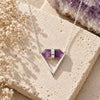 Amethyst Double Terminated Triangle Necklace featuring a faceted purple crystal set horizontally in a silver V-shaped pendant, displayed on natural beige stone with delicate dried flowers.