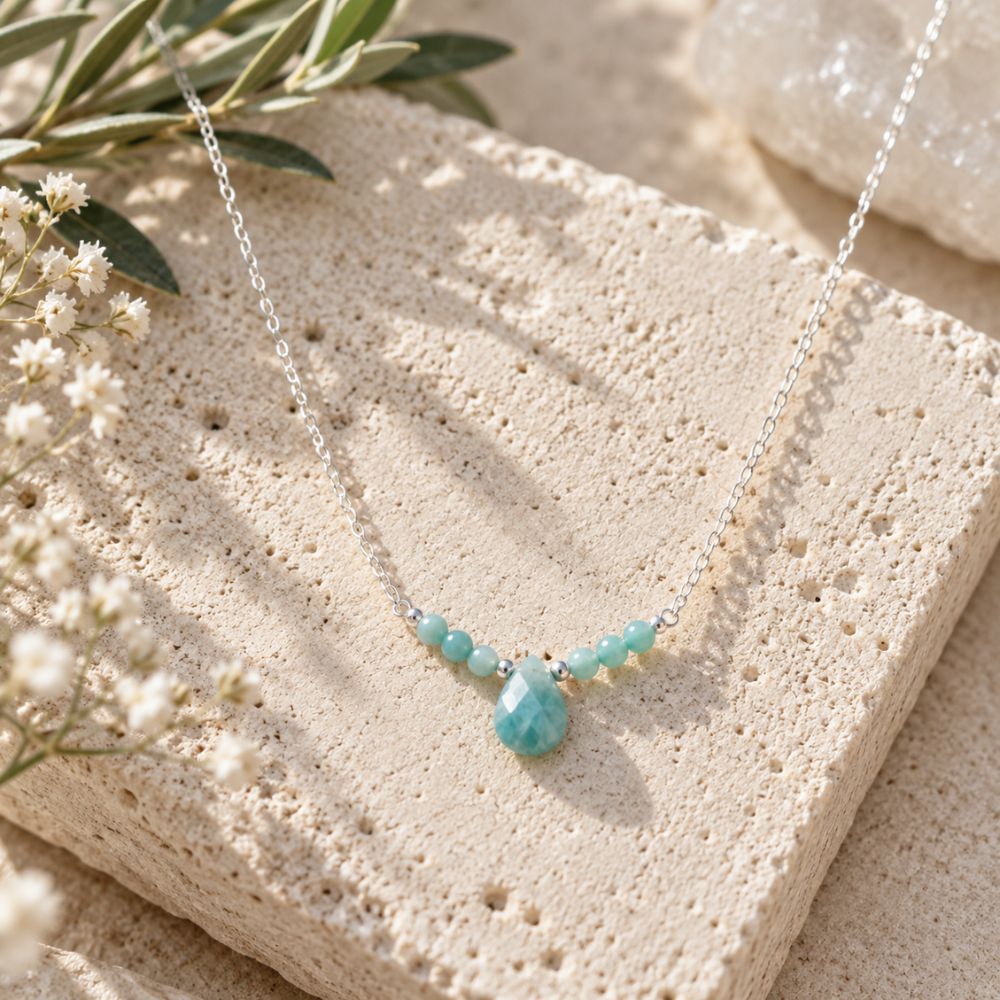 Amazonite Beaded Necklace featuring small aqua-green gemstone beads and a faceted teardrop pendant on a delicate silver chain, displayed on natural beige stone with soft greenery and white flowers.