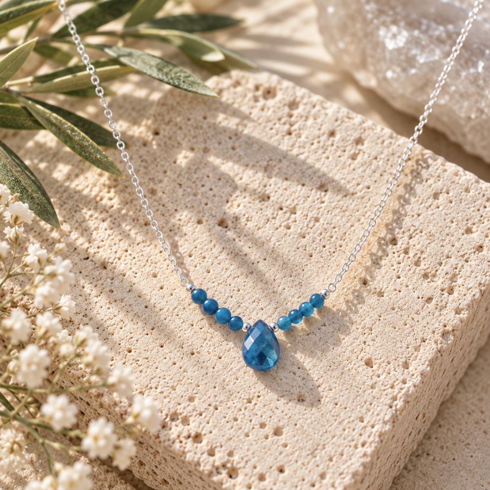 Blue Apatite Beaded Necklace featuring small blue gemstone beads, a faceted teardrop pendant, and a delicate silver chain, displayed on natural cream travertine with olive leaves and white flowers.