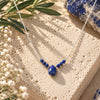 Lapis Lazuli Beaded Necklace featuring rich royal blue gemstone beads and a faceted teardrop pendant on a delicate silver chain, displayed on natural cream travertine with olive leaves and delicate white flowers.