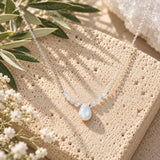 Moonstone beaded necklace featuring a faceted pear-shaped moonstone pendant, delicate white gemstone beads, and sterling silver chain, displayed on natural beige stone with olive branches and soft white flowers.